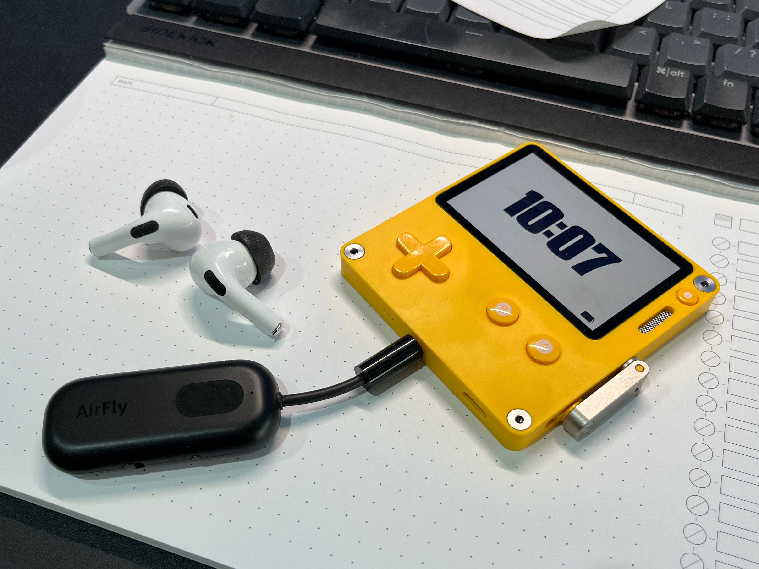 A PlayDate game console with an AirFly Pro bluetooth transmitter connected to its headphone port next to a pair of AirPods Pro earbuds